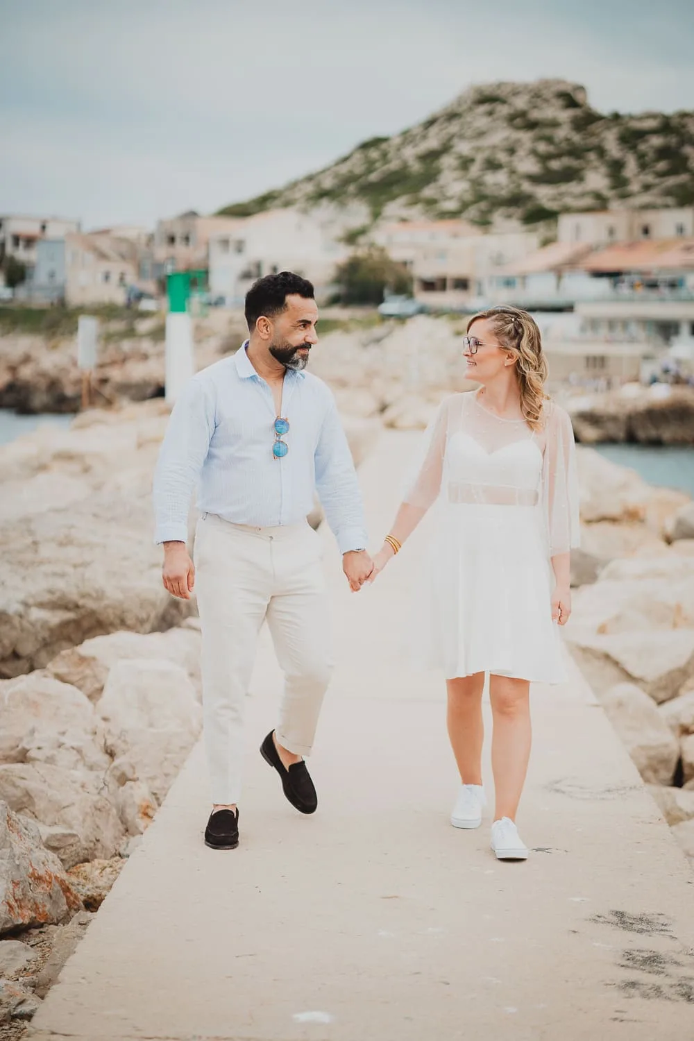 Couple de mariés marchant sur le quai des Goudes à Marseille, en bord de mer, au coucher du soleil. Photo de mariage dans un décor méditerranéen, entre bateaux, rochers.