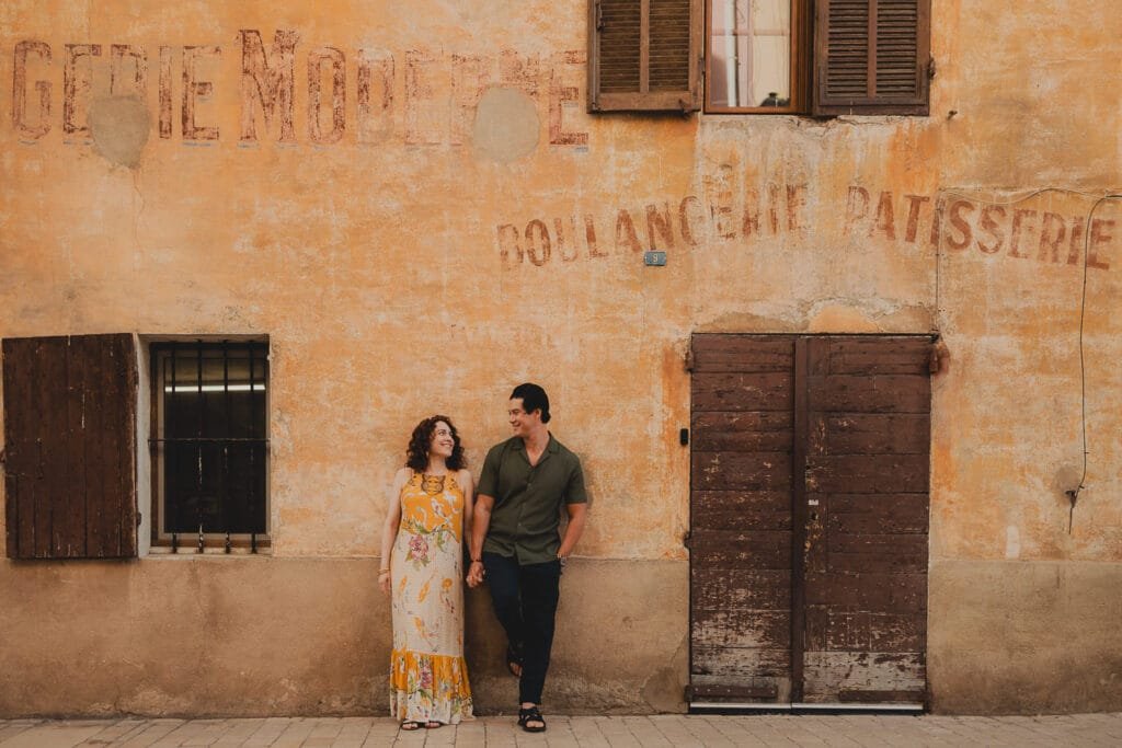 Couple posant devant une façade patinée dans le centre de Cassis, avec l’inscription « boulangerie » visible en arrière-plan